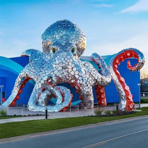 Escultura de Polvo Gigante envolvendo a entrada com texturas brilhantes de aço inoxidável